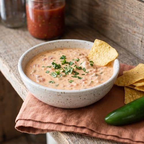 A speckled bowl of warm salsa and cream cheese dip topped with cilantro, with corn tortilla chips, a jalapeño and a jar of salsa on a wooden shelf