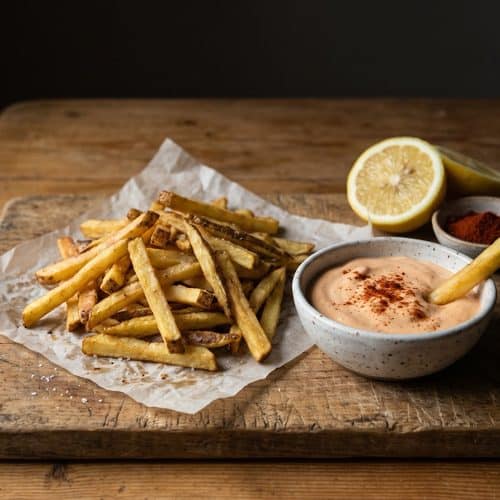 A pile of golden skin-on fries on parchment beside a speckled bowl of pale orange smoked paprika aioli dusted with paprika, with a halved lemon and a small dish of smoked paprika on a wooden board