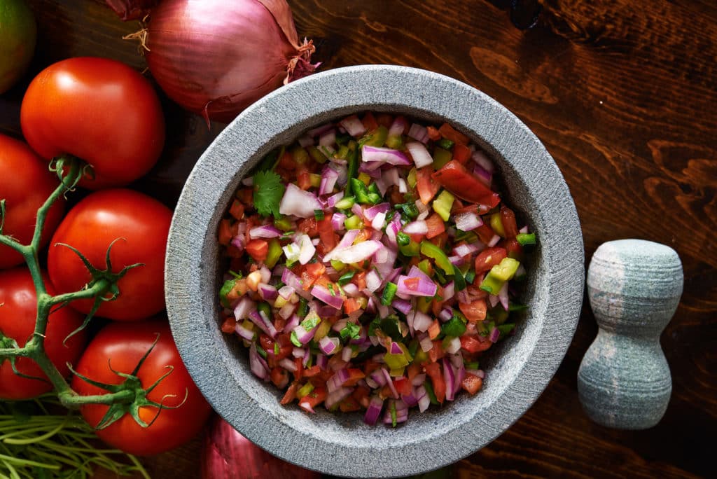 Fresh salsa of diced tomato, red onion and green chili in a stone molcajete, with whole tomatoes and a red onion beside it