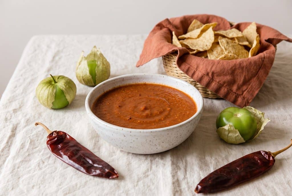 A speckled bowl of rusty red guajillo salsa with tortilla chips, dried guajillo chilies and fresh tomatillos on a cream tablecloth.