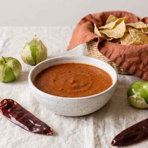 A speckled bowl of rusty red guajillo salsa with tortilla chips, dried guajillo chilies and fresh tomatillos on a cream tablecloth