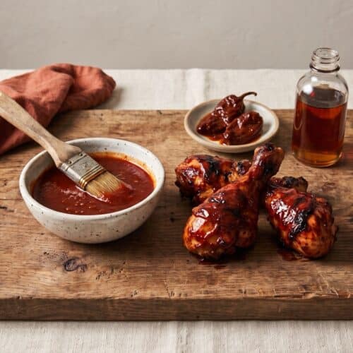 A speckled bowl of brick-red maple chipotle barbecue sauce with a basting brush, glazed grilled chicken drumsticks, two chipotles in adobo and a small bottle of maple syrup on a wooden board