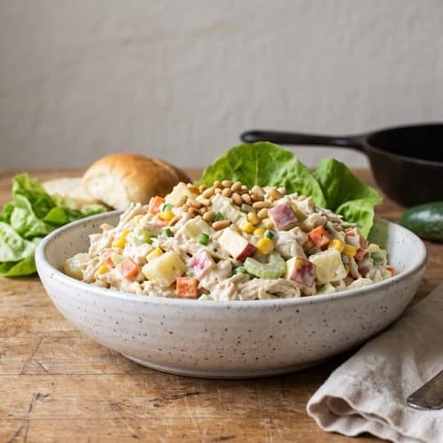 A speckled white bowl of chunky chicken salad with potato, carrot, corn, red apple, celery and diced jalapeño, topped with pine nuts, beside butter lettuce and a soft roll on a butcher-block counter