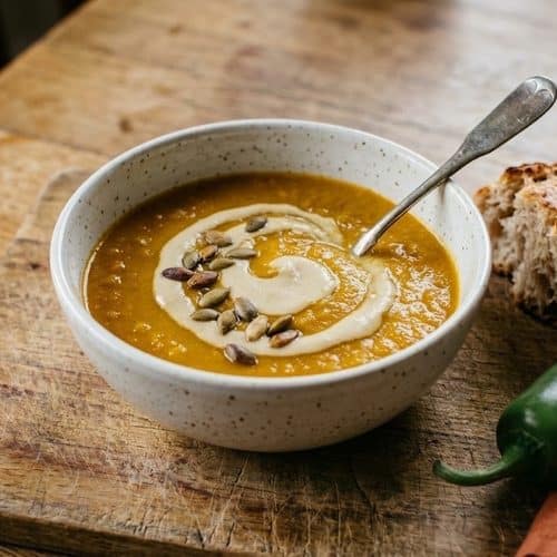 A speckled white bowl of spicy butternut squash soup with a swirl of cashew cream and toasted pepitas, beside crusty bread and a whole jalapeño on a worn cutting board