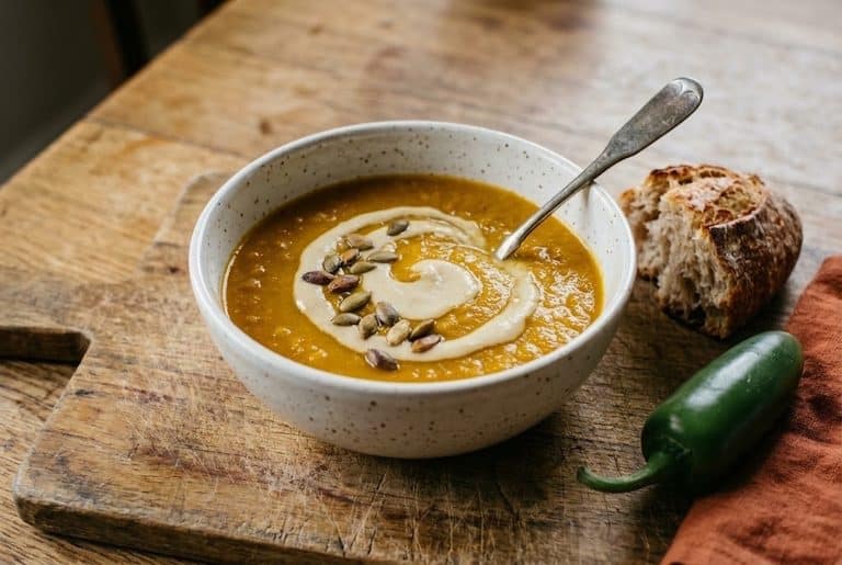 A speckled white bowl of spicy butternut squash soup with a swirl of cashew cream and toasted pepitas, beside crusty bread and a whole jalapeño on a worn cutting board