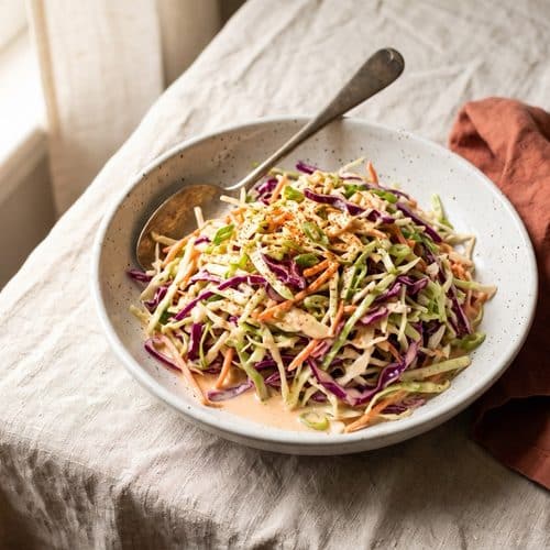 A small white bowl of creamy coleslaw with shredded cabbage and carrot on a gray wooden table