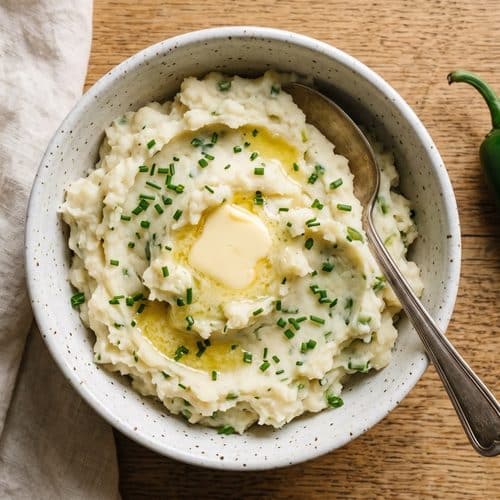 Overhead view of rustic jalapeƱo mashed potatoes in a speckled white bowl, topped with a melting pat of butter and chopped chives, with a spoon resting in the bowl and a whole jalapeƱo on an oak table