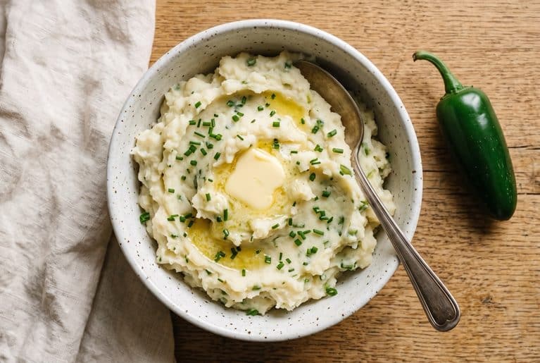 Overhead view of rustic jalapeño mashed potatoes in a speckled white bowl, topped with a melting pat of butter and chopped chives, with a spoon resting in the bowl and a whole jalapeño on an oak table