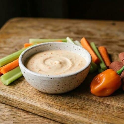 A speckled white bowl of creamy habanero ranch sauce with tiny bits of orange pepper, surrounded by celery and carrot sticks and a whole orange habanero on a worn cutting board