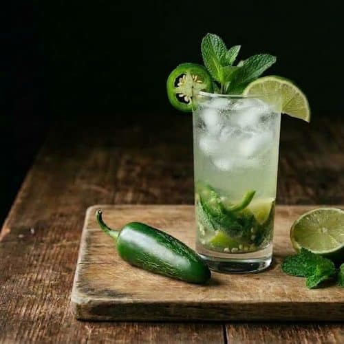 A tall jalapeƱo mojito with ice, muddled mint and lime, garnished with a jalapeƱo wheel and mint sprig, on a wooden board next to a whole jalapeƱo, half a lime and fresh mint