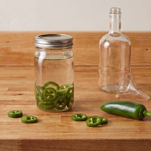 A mason jar of vodka with floating jalapeƱo rings beside an empty bottle, a funnel, a shot glass and a fresh jalapeƱo on a butcher-block counter