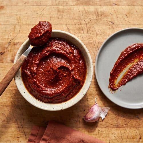 Overhead view of homemade aji panca paste in a speckled cream bowl on a butcher-block counter. A spoon holding a dollop of the thick brick-red paste rests across the bowl, and a swipe of paste is spread on a gray plate beside it, next to a garlic clove.