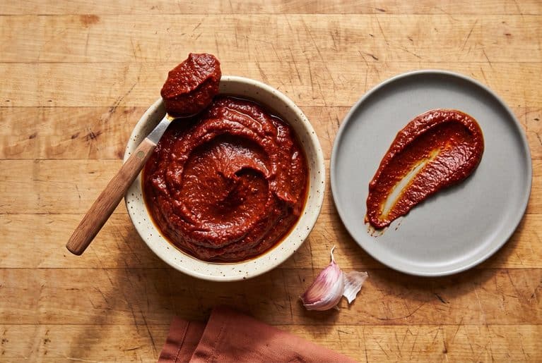 Overhead view of homemade aji panca paste in a speckled cream bowl on a butcher-block counter. A spoon holding a dollop of the thick brick-red paste rests across the bowl, and a swipe of paste is spread on a gray plate beside it, next to a garlic clove.