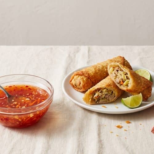 A small glass bowl of glossy sweet chili sauce flecked with chili, garlic and carrot, next to golden egg rolls and a lime wedge on cream linen