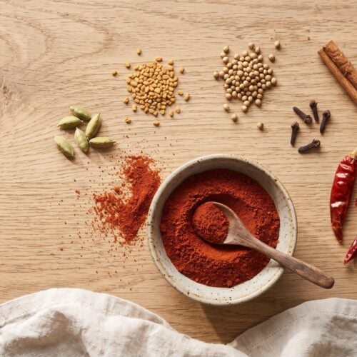 A speckled bowl of brick-red berbere spice mix with a wooden spoon, surrounded by cardamom pods, fenugreek and coriander seeds, cloves, a cinnamon stick and dried red chilies on an oak table