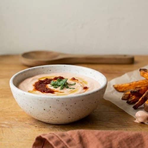 A bowl of pale pink harissa yogurt topped with a swirl of harissa, olive oil and mint, next to sweet potato fries on a butcher-block counter