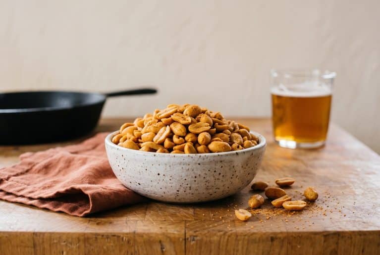A speckled white bowl of Old Bay peanuts on a butcher-block counter, with a few loose peanuts and seasoning spilled beside it and a glass of beer in the background.
