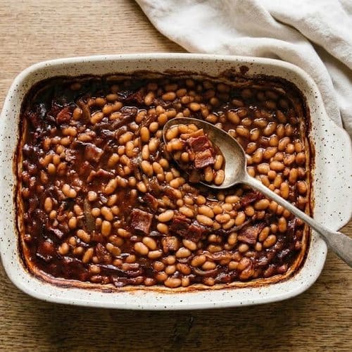 Overhead view of smoky and spicy baked beans with bacon in a speckled white baking dish, with a serving spoon resting in the thick, glossy sauce, on an oak table