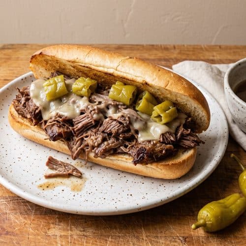 A toasted hoagie roll piled with shredded pepperoncini beef, melted provolone and whole pepperoncini, with a small bowl of juice for dipping on a butcher-block counter