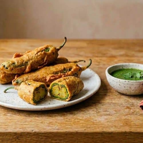 A speckled white plate of golden chilli pakoras with their green stems on, one cut open to show the spiced potato filling, beside a small bowl of green coriander chutney on a butcher-block counter