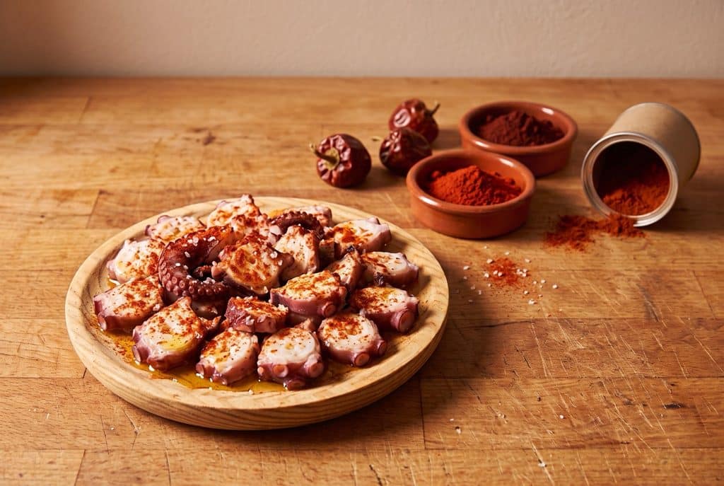 A wooden plate of sliced boiled octopus dusted with paprika and coarse salt, with two terracotta dishes of paprika, dried round red peppers and a tipped tin spilling paprika on a butcher-block counter