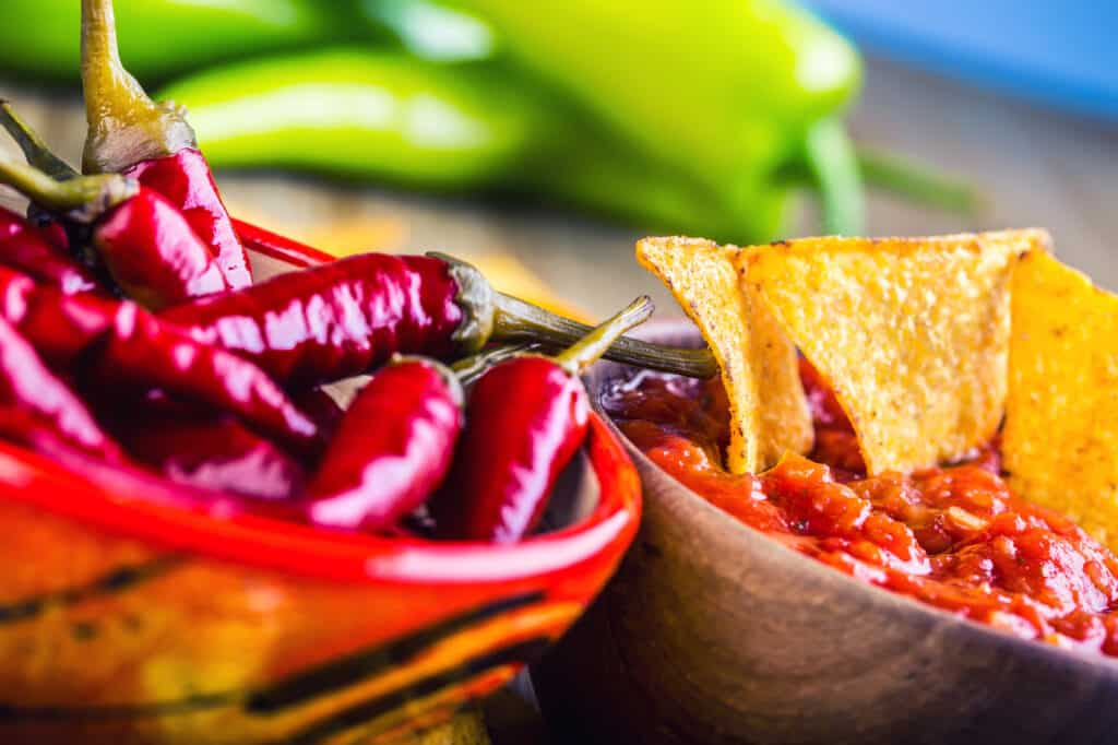 A bowl of red chili peppers beside a wooden bowl of salsa with tortilla chips, with long green peppers behind