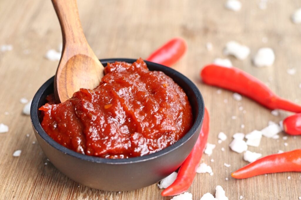 A small black bowl of thick red gochujang with a wooden spoon, surrounded by red chilies and coarse salt