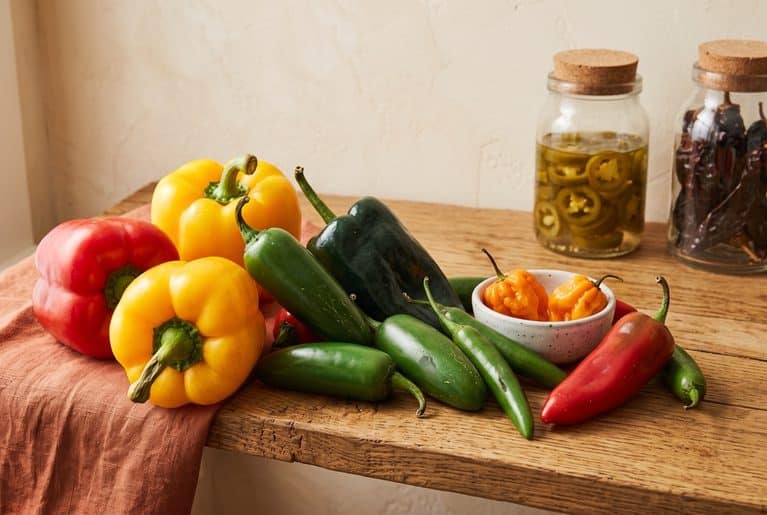 Red and yellow bell peppers, a dark green poblano, jalapeños, serranos and a red Fresno pepper on a rustic wooden shelf, with two orange habaneros in a small white bowl and jars of pickled jalapeños and dried chiles behind.
