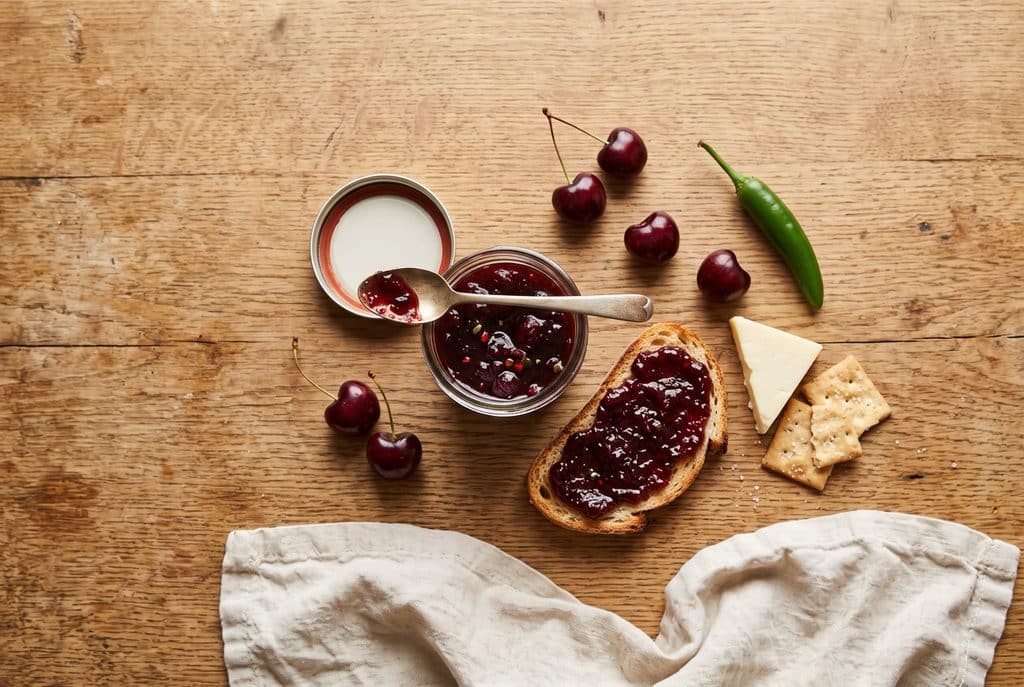A jar of spicy cherry jam with a spoon, toast spread with jam, white cheddar, crackers, fresh cherries and a serrano pepper on an oak table