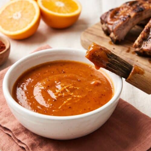 A bowl of orange chipotle sauce with a basting brush, halved oranges, chipotle powder and glazed pork ribs